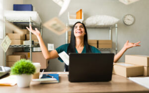 A woman sits at a desk with a laptop in a shipping area, joyfully throwing papers into the air with both hands raised in celebration. Cardboard boxes and packages are visible on shelving units in the background.